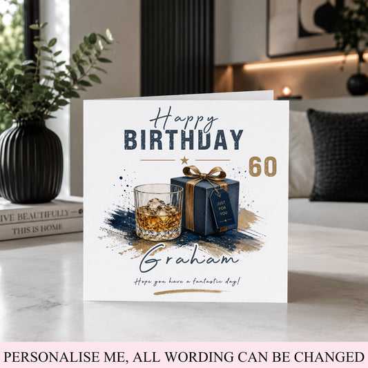 A personalized 60th birthday greeting card displayed on a marble tabletop in a modern living room setting. The white card features a dark blue and gold illustration of a whiskey glass and a gift box, with the customizable name 'Graham' at the bottom.