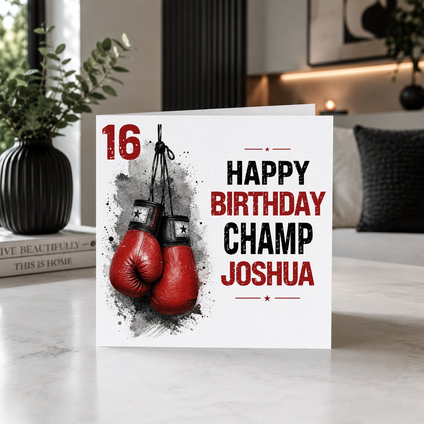 A personalized birthday card for a 16th birthday boxer, featuring red boxing gloves and the text 'HAPPY BIRTHDAY CHAMP JOSHUA' on a white card, styled on a marble tabletop in a modern living room setting.