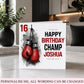 A personalized birthday card for a 16th birthday boxer, featuring a graphic of two red boxing gloves and the text 'HAPPY BIRTHDAY CHAMP JOSHUA' on a white card, displayed in a modern living room setting.