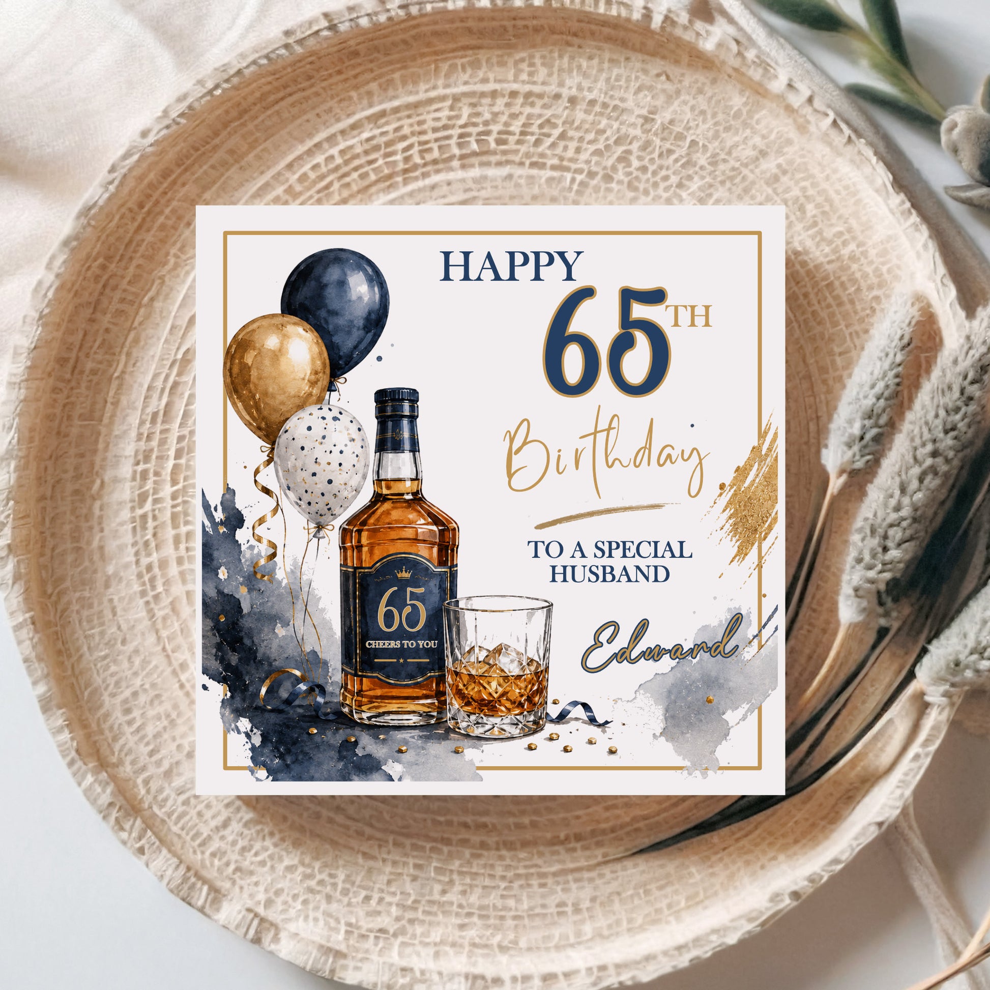 A square personalized 65th birthday card for a husband, styled flat-lay on a woven beige placemat. The card features a watercolor illustration of a whiskey bottle, a glass of amber liquor, balloons, and gold and navy text reading 'HAPPY 65th Birthday TO A SPECIAL HUSBAND' with the name 'Edward'.