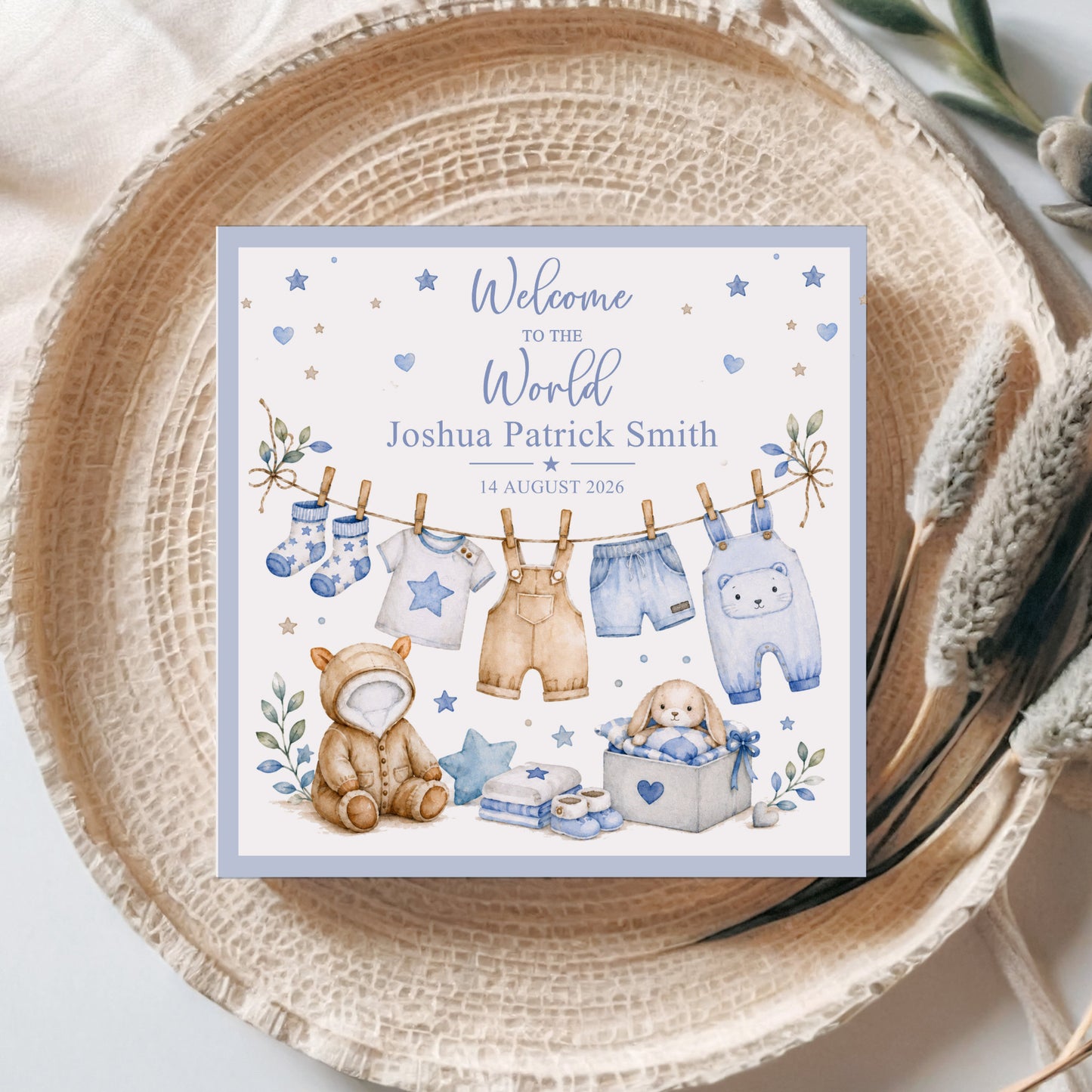 A personalized baby shower sign with a watercolor illustration of baby clothes hanging on a line, a teddy bear, and a bunny in a basket, placed on a woven placemat with dried grasses.