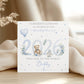 A personalized baby shower congratulations card for the birth of a boy, featuring watercolor-style illustrations of a teddy bear, balloons, flowers, and the year 2026. The card is displayed on a light wooden surface draped with soft beige fabric.