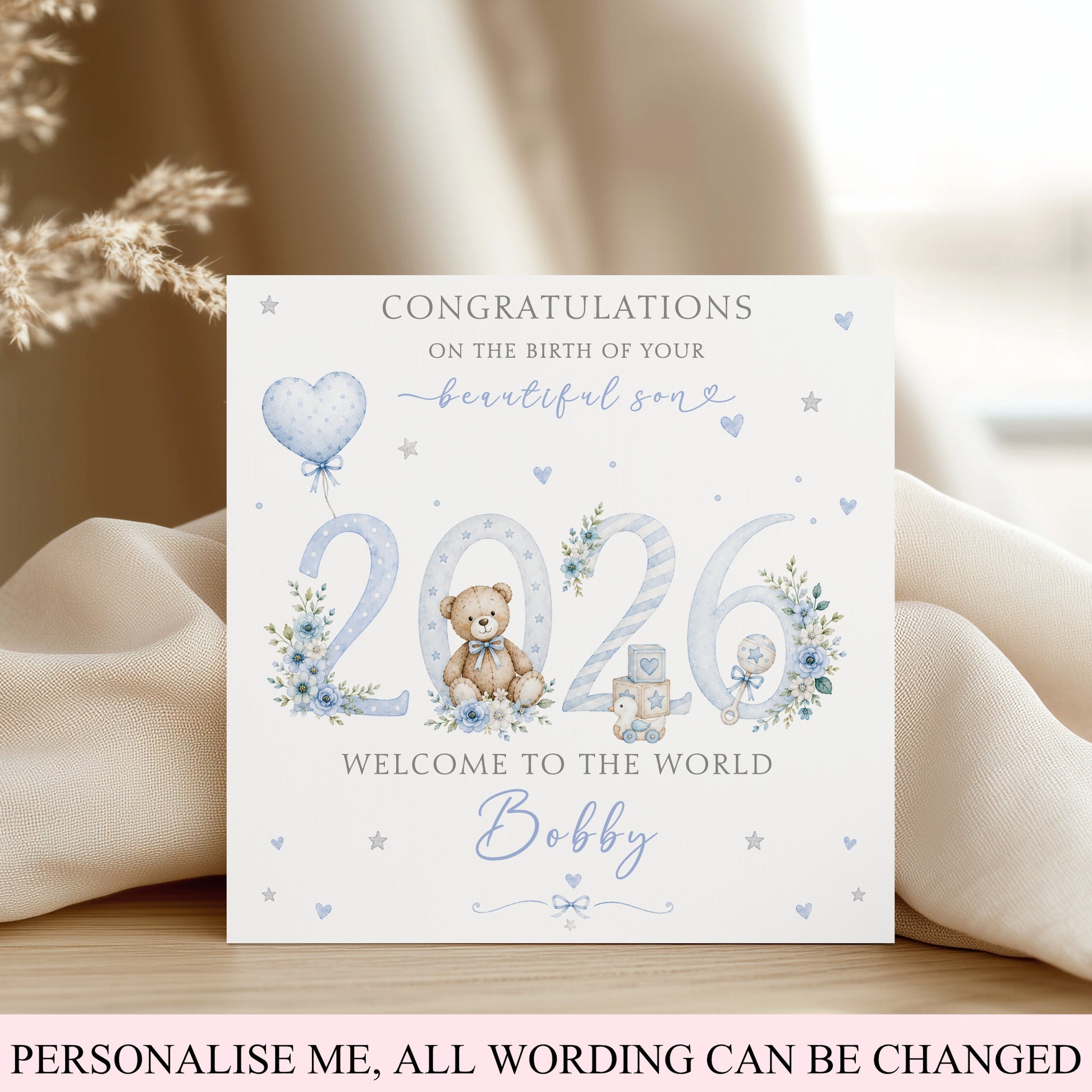 A personalized baby birth announcement card featuring a watercolor teddy bear, balloons, flowers, and the text 'Congratulations on the birth of your beautiful son 2026 Welcome to the world Bobby'. The card is displayed on a wooden surface draped with cream-colored fabric.