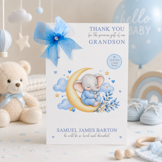 A white new baby grandson greeting card displayed upright in a soft blue and neutral nursery setting. The card features a sleeping baby elephant dressed in blue, resting on a golden crescent moon and surrounded by blue clouds, stars, hearts and delicate blue and cream flowers. The card reads “Thank You for the Precious Gift of Our Grandson” and includes personalised birth details for Samuel James Barton, followed by the message “He Will Be So Loved and Cherished.”