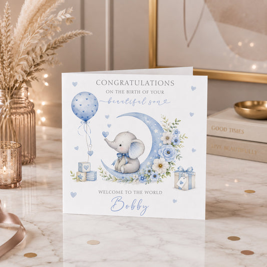 A personalized baby shower congratulations card featuring a watercolor illustration of a baby elephant on a crescent moon with blue flowers, balloons, and gifts. The card is displayed on a marble tabletop with decorative props like a gold vase, dried grass, and books.