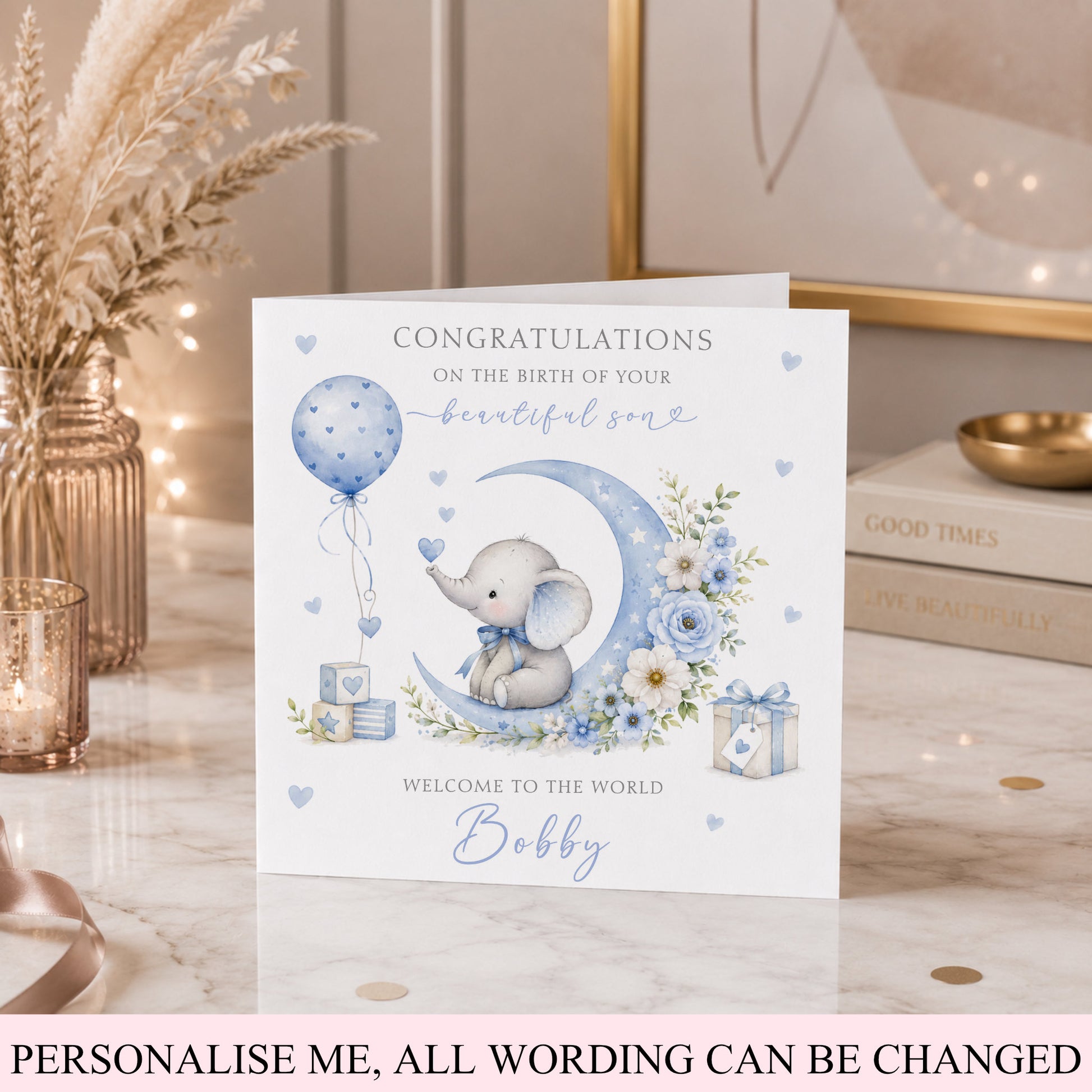 A personalized baby shower congratulations card featuring a watercolor illustration of a baby elephant on a crescent moon with blue flowers and balloons. The card is displayed on a marble tabletop with decorative dried grass, a candle, and books in the background.