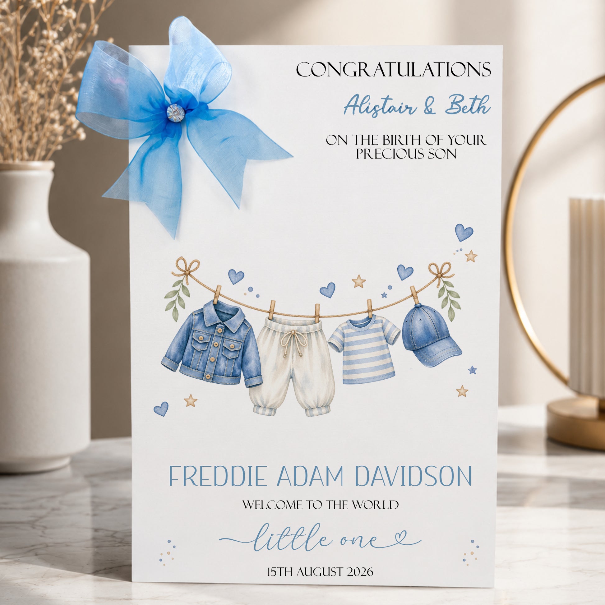 A white new baby boy greeting card displayed upright in a softly styled neutral setting. The card features a blue denim baby jacket, cream trousers, a blue and white striped top and a matching blue baby cap hanging from a decorative clothesline. Blue hearts, gold stars and delicate botanical details surround the clothing.