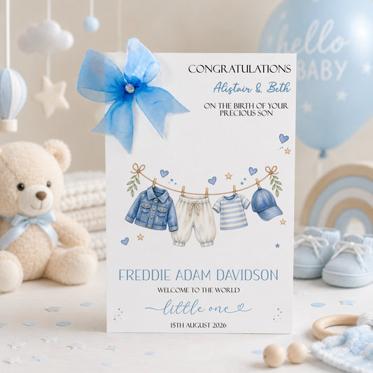 A white new baby boy greeting card displayed upright in a softly styled neutral setting. The card features a blue denim baby jacket, cream trousers, a blue and white striped top and a matching blue baby cap hanging from a decorative clothesline. Blue hearts, gold stars and delicate botanical details surround the clothing.