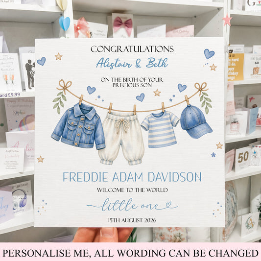 A personalized baby birth announcement card held by a hand, featuring an illustration of a baby's clothing hanging on a clothesline. The card is set against a background of retail shelves filled with other greeting cards.