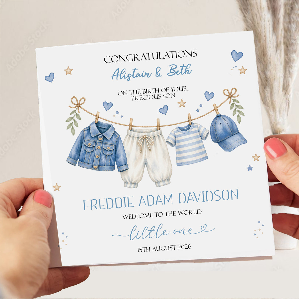 A personalized baby shower congratulations card held by two hands. The white card features a watercolor illustration of a baby boy's clothing set hanging on a clothesline, with custom text printed below.
