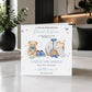 A personalized new baby congratulations card displayed on a marble countertop in a modern kitchen setting. The white card features watercolor illustrations of a teddy bear, a basket with a baby outfit, and blue shoes, with customizable text in blue and black.