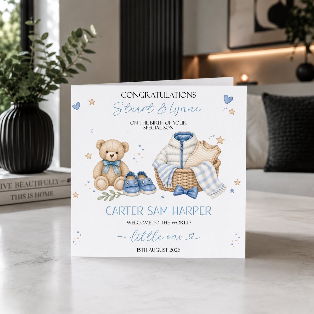 A personalized new baby congratulations card displayed on a marble countertop in a modern kitchen setting. The white card features watercolor illustrations of a teddy bear, a basket with a baby outfit, and blue shoes, with customizable text in blue and black.