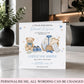 A personalized new baby congratulations card displayed on a marble countertop in a modern kitchen setting. The white card features watercolor illustrations of a teddy bear, a basket with a baby outfit, and blue shoes, with customizable text.