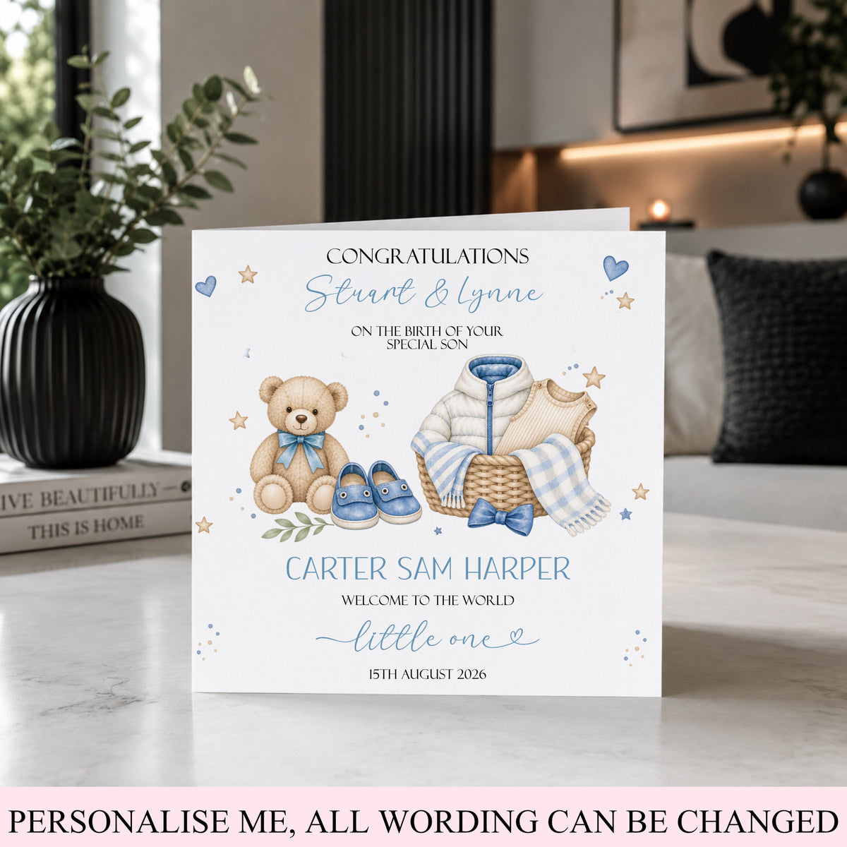 A personalized new baby congratulations card displayed on a marble countertop in a modern kitchen setting. The white card features watercolor illustrations of a teddy bear, a basket with a baby outfit, and blue shoes, with customizable text.