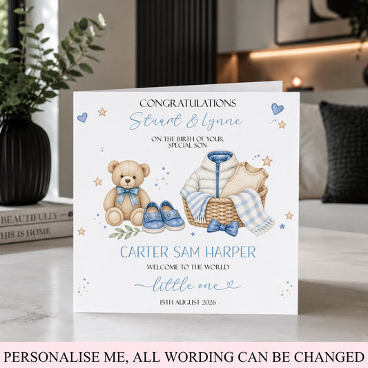 A personalized baby shower congratulations card displayed on a marble surface in a styled home setting. The card features watercolor illustrations of a teddy bear, a basket with baby clothes, and a pair of blue shoes, with customizable text.