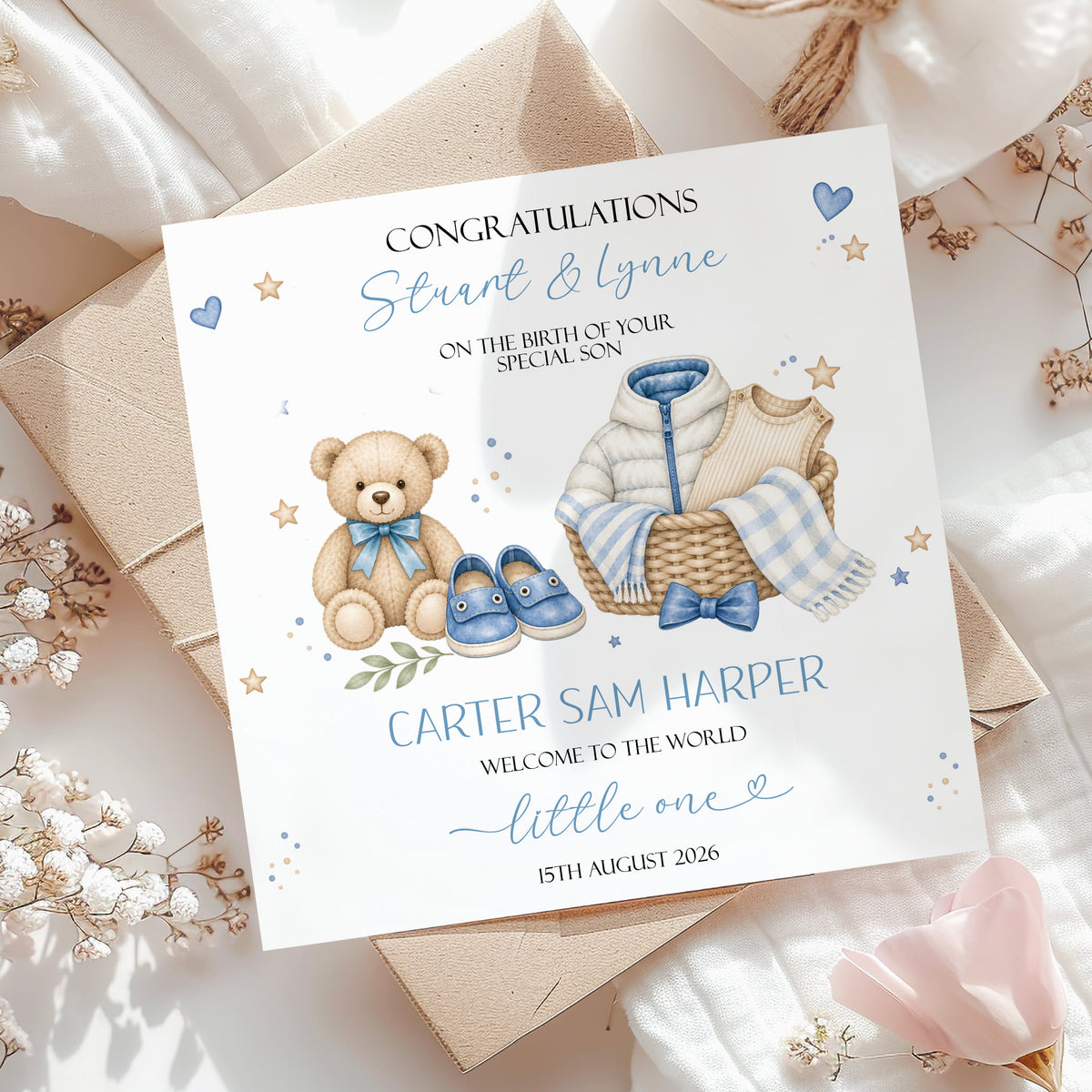 A personalized new baby congratulations card styled on top of brown kraft envelopes and white fabric with dried flowers. The card features a watercolor illustration of a teddy bear, baby clothes, and blue shoes, with customizable text reading 'Congratulations Stint & Lynne on the birth of your special son, Carter Sam Harper' and 'Welcome to the world, little one'.