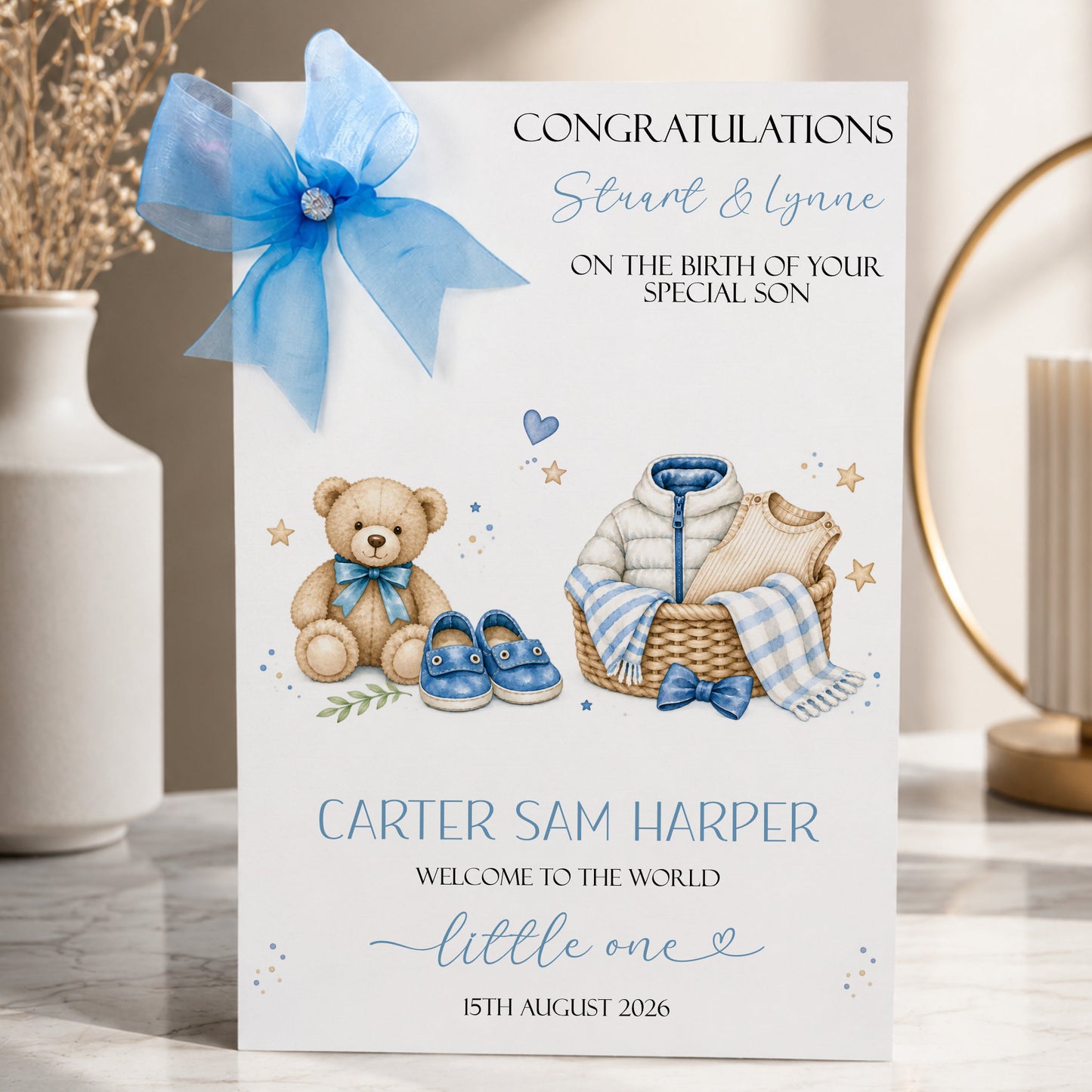 A white new baby boy greeting card displayed upright in a softly styled neutral setting. The card features a teddy bear wearing a blue bow, tiny blue baby shoes, a woven basket containing baby clothing and a blue and white blanket, surrounded by small stars and delicate botanical details. The card reads “Congratulations Stuart & Lynne” and “On the Birth of Your Special Son”, with “Carter Sam Harper”, “Welcome to the World Little One” and the date “15th August 2026” displayed below.