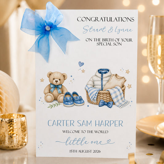 A white new baby boy greeting card displayed upright in a softly styled neutral setting. The card features a teddy bear wearing a blue bow, tiny blue baby shoes, a woven basket containing baby clothing and a blue and white blanket, surrounded by small stars and delicate botanical details. The card reads “Congratulations Stuart & Lynne” and “On the Birth of Your Special Son”, with “Carter Sam Harper”, “Welcome to the World Little One” and the date “15th August 2026” displayed below.