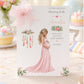A white baby shower greeting card displayed upright in a soft pastel pink celebration setting. The card features an elegant pregnant woman wearing a flowing pink dress, surrounded by delicate pink and green floral arrangements and a decorative feathered design. The card reads “To the Fabulous Mummy to Be” and “With Love on Your Baby Shower” and features the name Amelia in pink script. The surrounding scene includes soft pastel decorations, confetti and a beautifully styled baby shower setting.