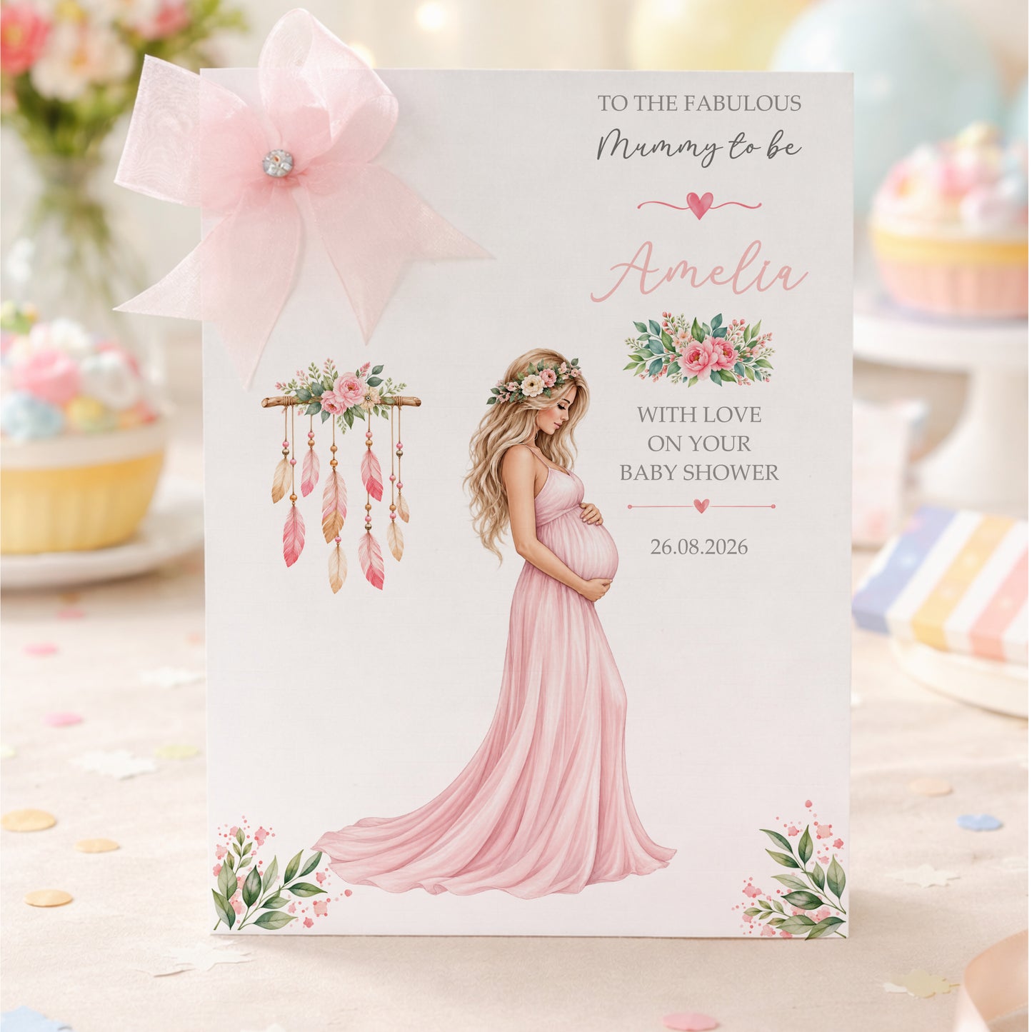 A white baby shower greeting card displayed upright in a soft pastel pink celebration setting. The card features an elegant pregnant woman wearing a flowing pink dress, surrounded by delicate pink and green floral arrangements and a decorative feathered design. The card reads “To the Fabulous Mummy to Be” and “With Love on Your Baby Shower” and features the name Amelia in pink script. The surrounding scene includes soft pastel decorations, confetti and a beautifully styled baby shower setting.