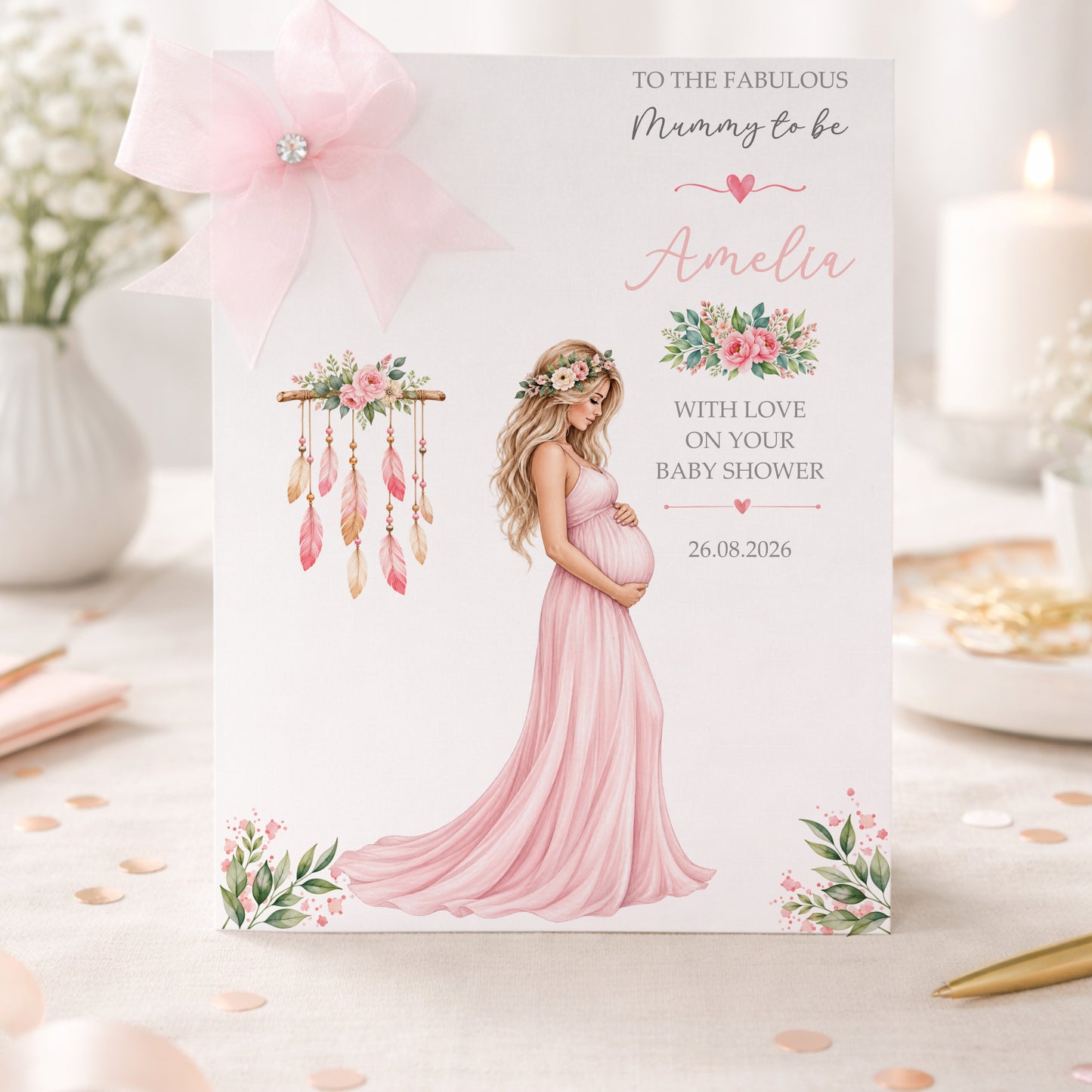 A white baby shower greeting card displayed upright in a soft pastel pink celebration setting. The card features an elegant pregnant woman wearing a flowing pink dress, surrounded by delicate pink and green floral arrangements and a decorative feathered design. The card reads “To the Fabulous Mummy to Be” and “With Love on Your Baby Shower” and features the name Amelia in pink script. The surrounding scene includes soft pastel decorations, confetti and a beautifully styled baby shower setting.