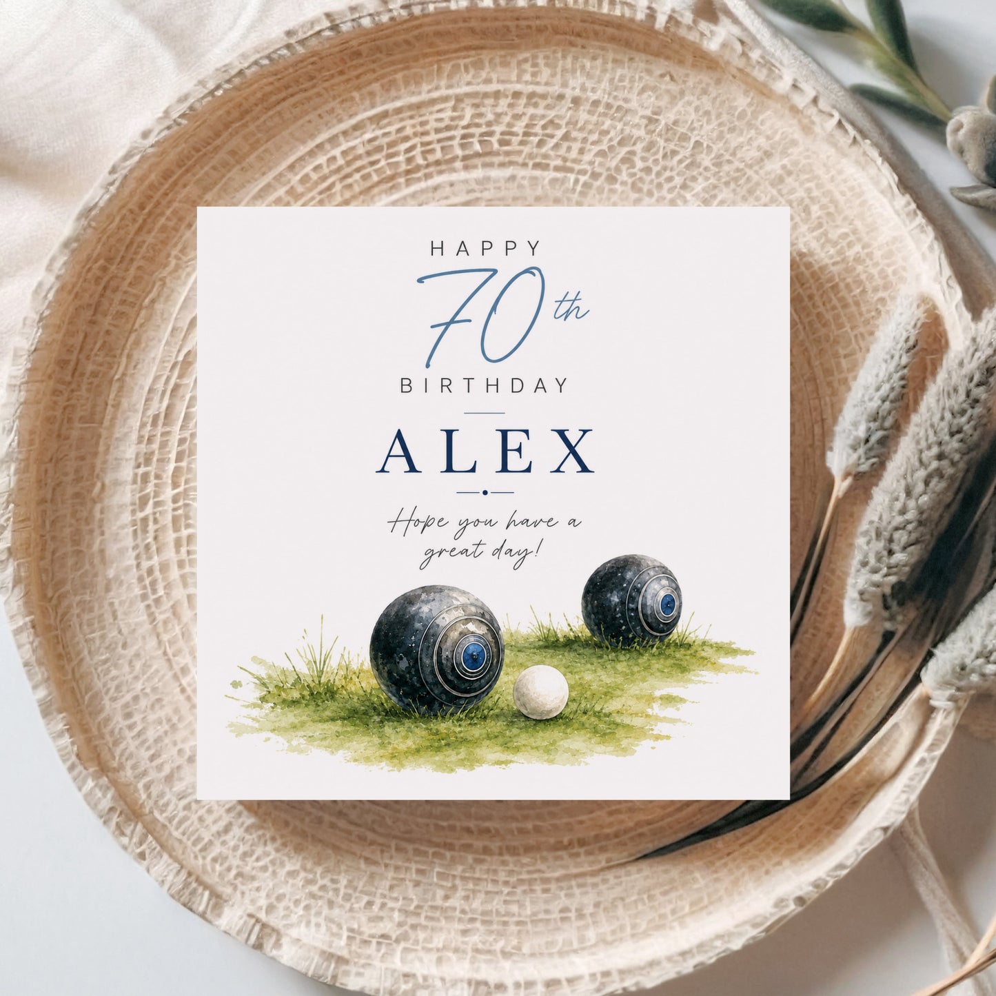 A square birthday card with a white background and blue text reading 'Happy 70th Birthday Alex' and 'Hope you have a great day!', styled on a woven beige plate with decorative dried grass and neutral fabric around it. The card artwork includes two black-and-blue croquet balls on green grass.