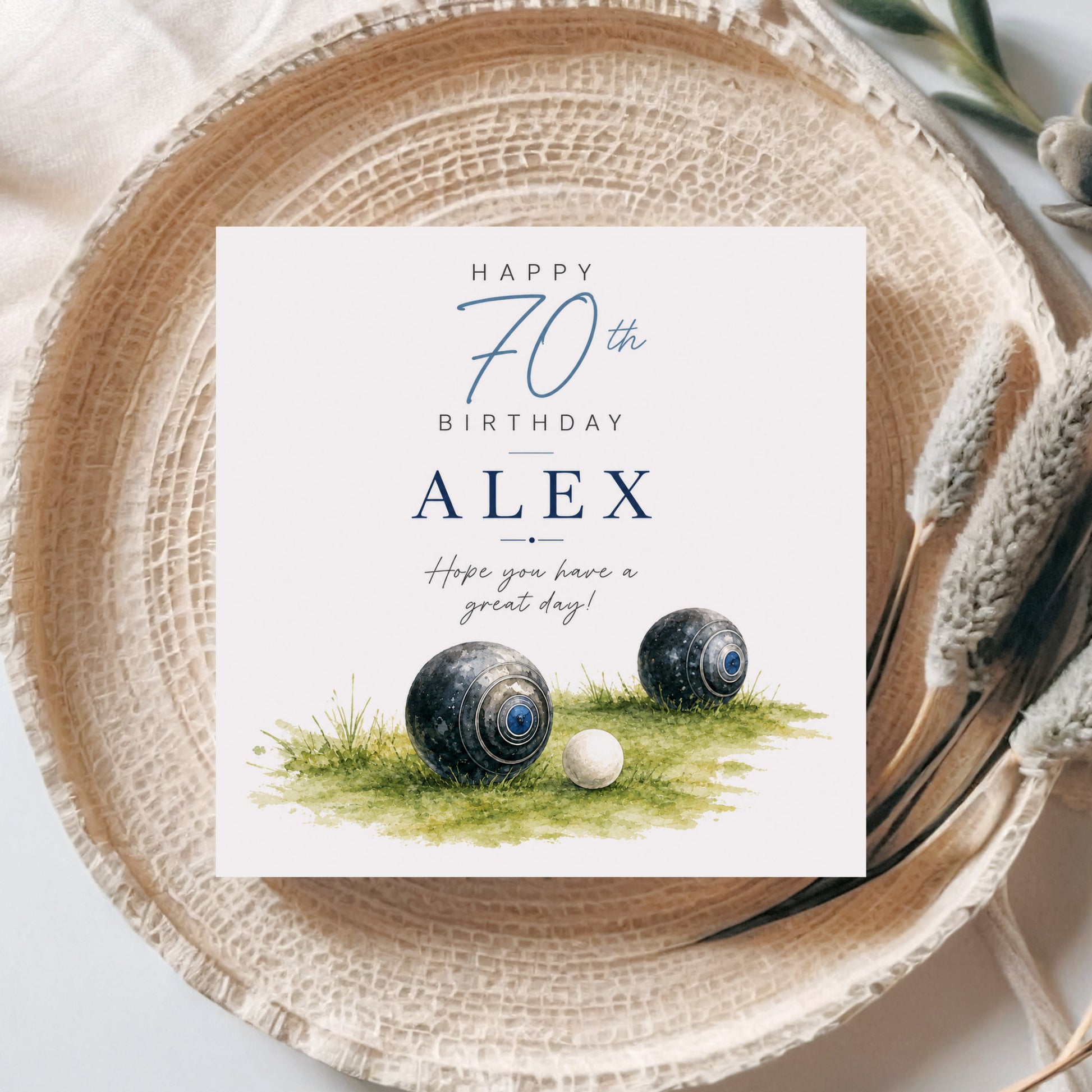 A square birthday card with a white background and blue text reading 'Happy 70th Birthday Alex' and 'Hope you have a great day!', styled on a woven beige plate with decorative dried grass and neutral fabric around it. The card artwork includes two black-and-blue croquet balls on green grass.