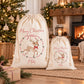 Two cream-colored drawstring Christmas gift bags with personalized designs, one large and one small, displayed in a cozy holiday living room setting with a fireplace, Christmas tree, and wrapped gift.