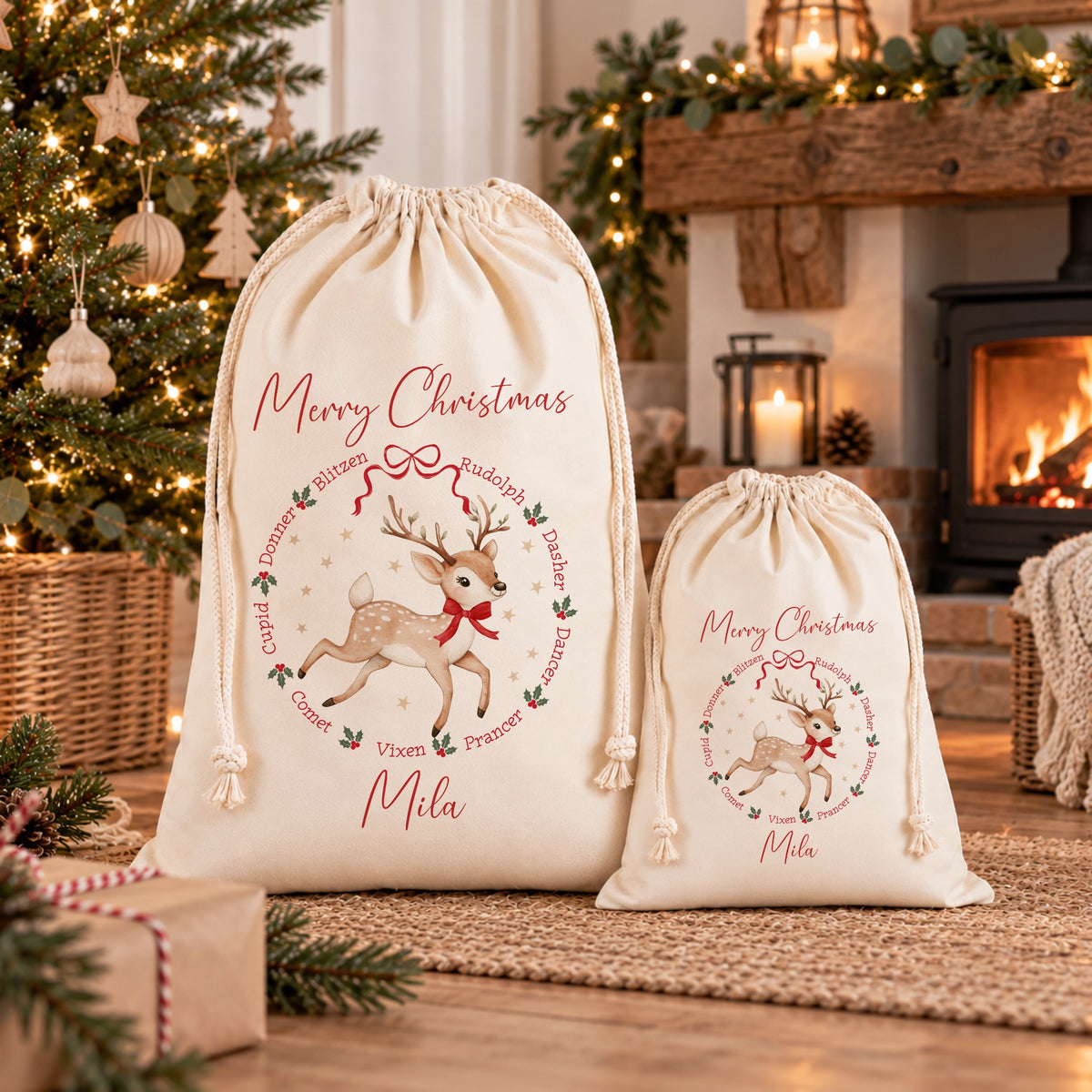 Two cream-colored drawstring Christmas gift bags with personalized designs, one large and one small, displayed in a cozy holiday living room setting with a fireplace, Christmas tree, and wrapped gift.
