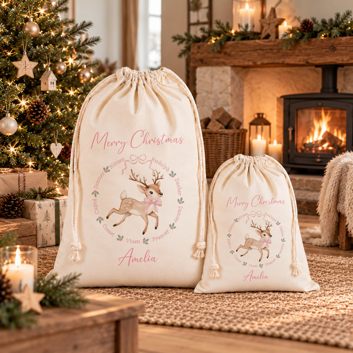 Two cream-colored personalized Christmas drawstring gift bags displayed in a cozy living room setting with a fireplace, Christmas tree, and wrapped gifts. The larger bag features a graphic of a reindeer inside a wreath with the text 'Merry Christmas' and the name 'Amelia' printed below.
