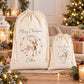 Two cream-colored canvas drawstring gift bags with Christmas deer illustrations and 'Merry Christmas Callum' text, displayed on a wooden table in a cozy, warmly lit living room setting with wrapped presents and a decorated Christmas tree in the background.