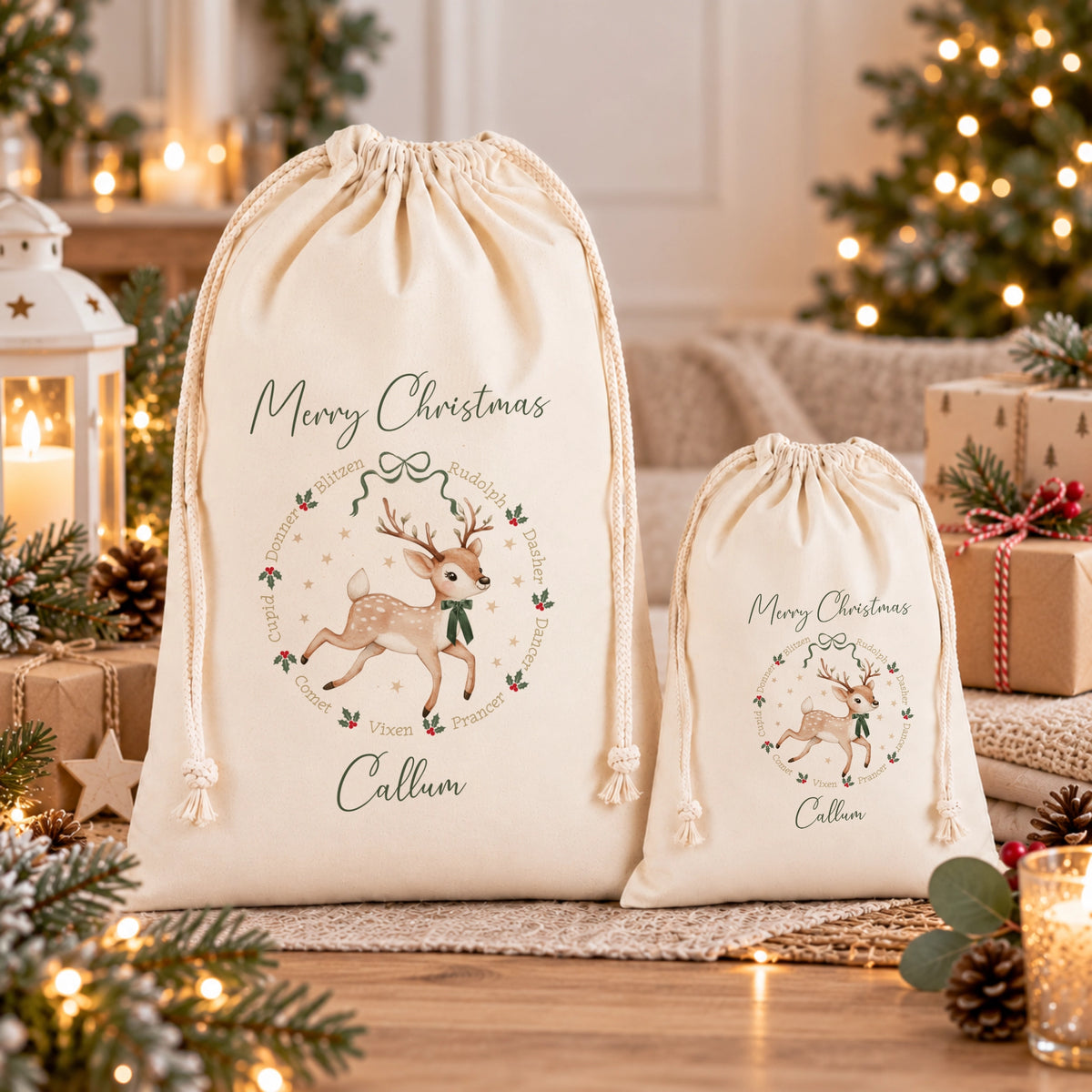 Two cream-colored drawstring Christmas gift bags with personalized names and a festive deer illustration, displayed on a wooden surface with a cozy, warmly lit Christmas background.