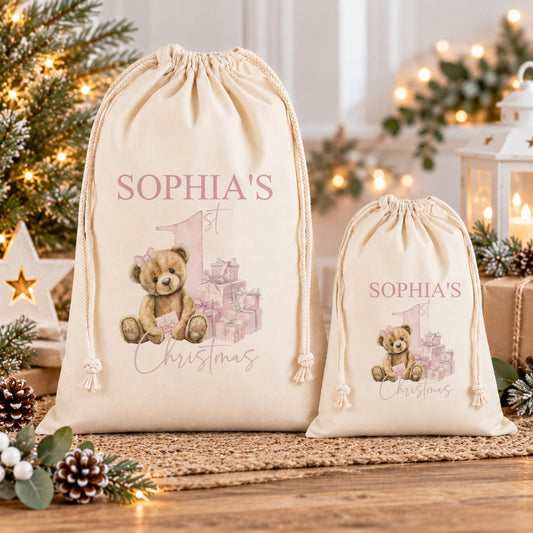 two white drawstring bags with a teddy bear design and the text "Sophia's 1st Christmas" printed on them. The bags are placed on a wooden surface, with a Christmas tree and other holiday decorations visible in the background.