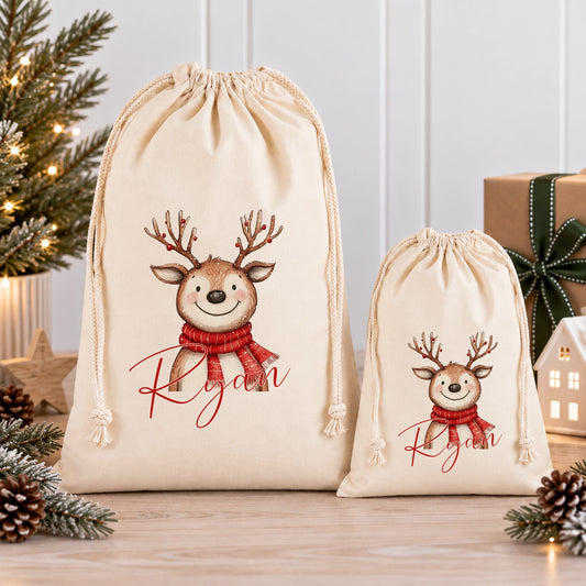 Two beige drawstring bags with a picture of a smiling deer wearing a red scarf and the name "Ryan" printed on them, placed on a wooden surface with pine cones and a small Christmas tree in the background.